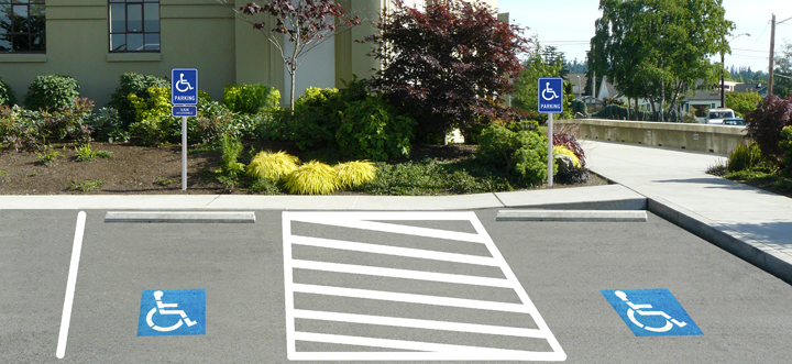 A well-marked accessible parking area with two wide parking bays featuring wheelchair symbols and crosshatch zones for extra space, located directly in front of a building entrance with a curb ramp nearby and signage indicating disability access.